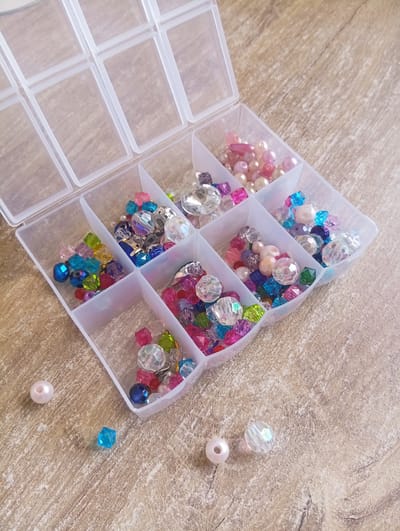This image depicts a clear plastic bead organizer with multiple compartments, filled with various colorful beads. The organizer is open, and the beads are predominantly in shades of pink, blue, green, clear, and purple. Some of the beads are faceted and transparent, while others are opaque pearls. A few individual beads, including a pink pearl, a blue faceted bead, and a clear faceted bead, are scattered on the wooden surface next to the organizer. The background is a light brown wooden surface with visible grain. There is no indication of a specific location in Hrodna, Belarus. The scene is static and shows a collection of crafting materials.