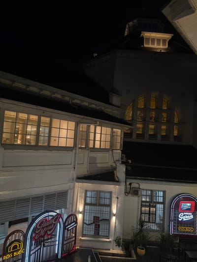 This image, taken at night in Medan, Indonesia, captures the exterior of a building with a retro aesthetic. The scene is illuminated by streetlights and the glow from interior lights filtering through windows. Neon signs are a prominent feature, with one large sign displaying "Gudang Garam" in red and white neon, accompanied by smaller signs reading "POS BLOC" in yellow neon. These signs suggest a commercial establishment, possibly a restaurant or cafe, given the "POS BLOC" text which likely refers to a public space or building.
The architecture features a mix of old and new elements. On the right side, a white building with multiple windows, some with distinctive arched stained-glass patterns, is visible. The windows on the lower floors are mostly unadorned, with one showing visible interior lighting. Air conditioning units are mounted on the exterior walls. To the left, another part of the building appears older, with a light-colored facade and prominent shutters or grilles on some windows. The overall impression is that of a lively urban environment at night, with the neon lights adding a vibrant touch to the architecture. No people are visible in the frame, and the weather appears clear, as indicated by the sharp, defined lighting. The orientation of the image is rotated, suggesting it was taken with the camera held sideways.