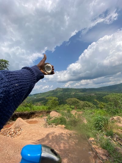 The image shows a person's arm reaching out towards the sky, holding a halved coconut. The person is wearing a dark blue knitted sweater. The scene is set on a rocky cliff overlooking a lush, green valley with rolling hills in the distance. The sky is filled with dramatic, white clouds against a bright blue backdrop. The sun is shining, suggesting it is daytime. The overall impression is one of freedom and connection with nature. The location cues, such as the vegetation and rock formations, are consistent with a tropical or subtropical region. The image appears to be taken in Amaigbo, Nigeria.