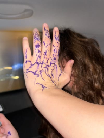 A person with brown, wavy hair is resting their head on their left hand. The left hand and wrist have purple markings resembling abstract designs or doodles. The person's right hand, with a purple dot on it, is visible in the lower right corner of the image. The background includes a grey wall or door with a handle, and a reflective surface, potentially a countertop. No information is available in the image to indicate the location is Rishon LeZion, Israel.