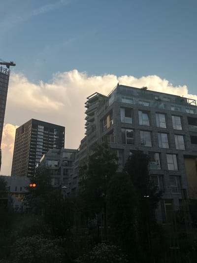 The image depicts a view of modern apartment buildings in Amsterdam, Netherlands, under a partially cloudy sky. In the foreground, dense foliage partially obscures the lower portions of the buildings. A crane is visible in the upper left corner, indicating ongoing construction. The buildings themselves are contemporary in design, with multiple stories, large windows, and balconies. One building on the left is a tall, dark structure with a grid-like pattern of windows. Adjacent to it is a lighter-colored building with a more complex, stepped facade. A prominent building in the center right features a grey brick exterior and glass railings on its balconies. Two round, orange lights are visible on a lower building in the midground, positioned between trees. The overall scene suggests an urban environment with a mix of completed and under-construction residential properties.