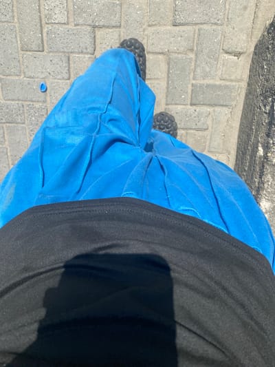 A street-level view shows a person standing on a gray brick sidewalk. The person is wearing blue pants with a textured, layered design, and a black shirt. The person's feet are clad in dark-colored sandals. A small, blue object lies on the sidewalk near the pants. The background is a concrete brick surface. The location is the city of Iranla, Nigeria.