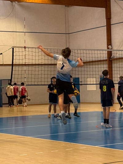 The image displays an indoor volleyball court within a gymnasium in Sennecey-le-Grand, France. A male athlete, wearing a light blue and white jersey with the number "7" on the back and black shorts, is captured in mid-air above the net, with his right arm drawn back in preparation for a spike. He wears light grey knee pads.
A black volleyball net with a white top tape stretches across the court, and a red and white striped antenna marks the net boundary on the left side. On the same side of the net as the jumping player, two other male players are visible; one wears a dark jersey with the number "7" and holds a yellow volleyball. On the opposite side of the net, a male player in a dark blue jersey with "SUC" and the number "8" printed on the back stands facing away from the camera.
In the far left background, four additional individuals are present off-court, near a light-colored wall. Two wear red jerseys with numbers "13" and "2," respectively, while two others wear hoodies. On the far right, a partially visible adult male in a dark tracksuit is seen walking. The gymnasium floor features a wooden perimeter surrounding a prominent blue playing area marked with white lines. The walls are light-colored with a single horizontal blue stripe, and wooden beams support the ceiling structure. Blue doors are visible in the background.