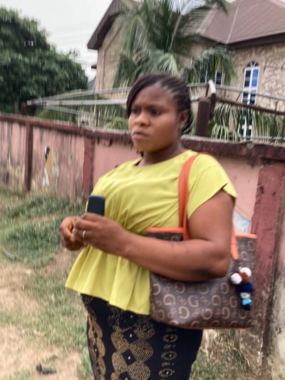 A woman is depicted standing outdoors in Aba, Abia, Nigeria, on an overcast day. She appears to be in her late 20s to early 30s, with dark skin and dark braided hair. She wears a light yellow, short-sleeved top with a pleated hem and a dark skirt featuring a gold geometric pattern. A silver-colored ring is visible on her left ring finger. She holds a dark-colored feature phone in her right hand. A brown shoulder bag with a repeating 'G' logo and orange trim hangs from her left shoulder, adorned with a small, multi-colored keychain. Her gaze is directed towards the left side of the frame, with a neutral expression. In the background, a faded reddish-pink wall with faint artwork is visible. Above the wall, a metal fence structure precedes a two-story building with a brown roof and arched, blue-tinted windows. Lush green foliage, including palm fronds, is present throughout the background. The ground in the foreground is unpaved earth with sparse vegetation.