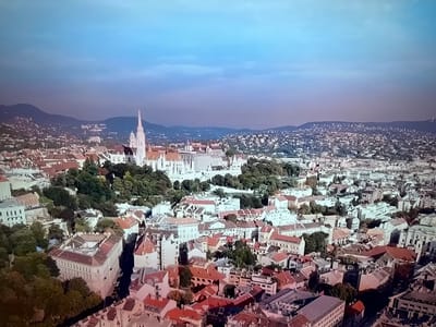 This is an aerial, high-angle, panoramic shot of a densely populated city, possibly Budapest, Hungary, given the prominent white Gothic-style church with a tall spire and red-tiled roofs on its left. The city features a mix of architectural styles, with many buildings constructed from light-colored stone or plaster. Some structures are multi-story residential buildings, while others appear to be larger public or religious buildings. Trees are interspersed throughout the cityscape, particularly around the church. In the background, rolling hills covered in more dense urban development rise against a hazy sky, transitioning from a deep blue at the top to a lighter, purplish hue near the horizon. The image does not depict the city of Ləcət, Azerbaijan. There are no people visible in the scene.