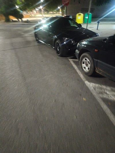 The image depicts an outdoor scene at night, likely in the city of Harmanli, Bulgaria, showing two parked cars on a street. In the foreground, the right side of a dark-colored car is visible, with its front wheel and part of the chassis in view. White parking lines are painted on the asphalt. Further back, a sleek, black sports car is parked, its front end and passenger side clearly visible. The sports car has shiny black rims. Beyond the cars, there are trees with illuminated foliage suggesting streetlights. In the background, two colorful trash bins, one yellow and one green, stand next to a blue bollard. A red "STOP" sign is also visible on a pole behind the sports car. The overall lighting indicates nighttime, with artificial light sources creating reflections and highlights on the vehicles and pavement.