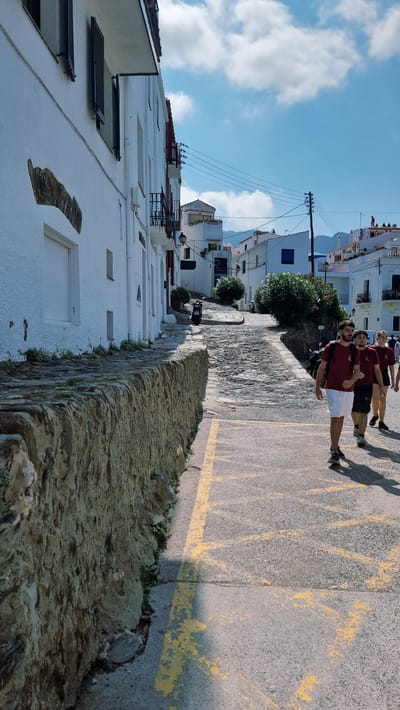 A group of four young men are walking down a narrow street in Cadaqués, Spain. They are dressed casually, with two of them wearing maroon t-shirts and white shorts, and the other two in similar attire. The street is paved with stone and lined with whitewashed buildings. The day appears to be sunny with a clear blue sky and some scattered clouds. The sun casts long shadows from the men onto the road, suggesting it is late afternoon. The buildings are typical of Mediterranean architecture, with some featuring blue shutters. There are power lines visible overhead. The scene conveys a sense of leisure and exploration as the men walk together.