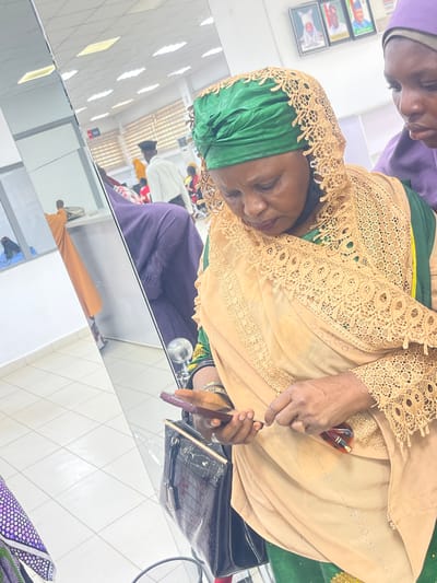 An indoor scene in Zaria, Nigeria, features a woman, likely middle-aged, positioned centrally on the right, looking downwards at a dark-colored smartphone held in both hands. She wears a green headwrap and a beige lace-edged veil over a green garment. Her left wrist displays multiple silver-colored bangles. A pair of brown-framed eyeglasses is held in her right hand near the phone. A dark brown, crocodile-patterned handbag with a gold-colored clasp is visible below her hands. The upper right corner shows the partial view of another woman, wearing a purple head covering, observing the main subject. The left side of the image is dominated by a large mirror, reflecting an interior space with white walls, a light-colored tiled floor, and several individuals, including one in an orange garment and others further back. On the wall in the upper right background, three framed portraits of men in traditional attire are mounted. The environment is brightly illuminated by overhead artificial lighting.