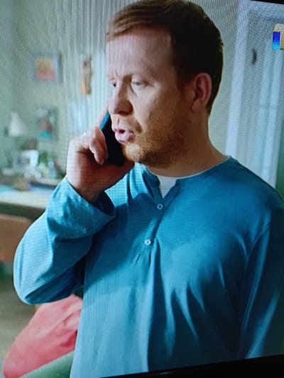 The media file shows a medium shot of a fair-skinned man with red hair and a beard, who appears to be in his late 30s or early 40s. He is wearing a light blue long-sleeved shirt with a white undershirt visible at the collar and a Henley neckline with two buttons. The man is holding a dark-colored mobile phone to his left ear and speaking into it, with his mouth slightly open. His expression is somewhat strained or concerned.

The background is blurred, suggesting an indoor setting. On the left side, there is a wall with what appears to be framed pictures or artwork. A desk or table with some items on it is also visible. The lighting suggests it is daytime, though the exact time is not clear. The overall mood conveyed is one of a private conversation, possibly a serious one, given the man's facial expression. There is no visible text in the image. The context provided places this scene in Shymkent, Kazakhstan.