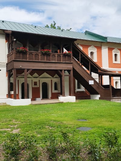 The image depicts the exterior of the Church of Elijah the Prophet in Yaroslavl, Russia, specifically showing its wooden porch and outdoor staircase. The building's facade is painted in alternating bands of orange and white. A prominent wooden porch with decorative railings and flower boxes is visible on the upper level, accessible by a wooden staircase. The staircase is dark brown and has a protective roof over its upper section. The ground in front of the building is covered in green grass. The sky above is partly cloudy.