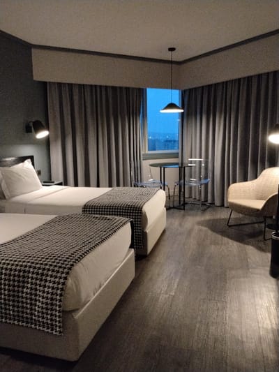 The image displays the interior of a modern hotel room in Porto, Portugal, depicted at twilight or nighttime. No individuals are present. The room contains two parallel single beds, each made with white linens and accented by a black and white houndstooth pattern bed runner. A dark wall-mounted reading lamp, illuminated, is positioned above the bed on the left. Between the beds, a small dark nightstand is visible. A large window, flanked by drawn grey pleated curtains, dominates the background, offering a view of a dark blue sky with indistinct city lights in the distance. A black conical pendant lamp hangs illuminated in front of the window. Adjacent to the window, a small round dark table is paired with two transparent, modern chairs. To the right, a light beige upholstered armchair with dark metal legs is situated, with a partially visible dark cylindrical object next to it. The floor is covered in dark grey wood-effect laminate. The walls are dark grey, topped by a white ceiling with a grey border.