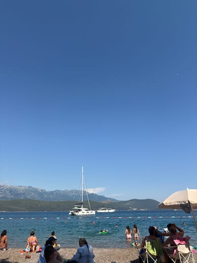In Radovići, Montenegro, a serene beach scene unfolds under a clear blue sky. The foreground is occupied by a diverse group of people enjoying the pebbled shoreline and the tranquil sea. Some are seated in beach chairs, others are sunbathing on towels, and a few are wading into the cool, clear water. The middle ground features several individuals swimming and playing in the sea, with a bright green inflatable toy visible. Further out, a majestic white catamaran with multiple sails is anchored, and a sleek white motor yacht is also present. The background is dominated by lush green hills that rise to meet imposing, rugged mountains under a vast expanse of sky. The image captures a leisurely summer day at the coast, with the natural beauty of Montenegro providing a stunning backdrop.