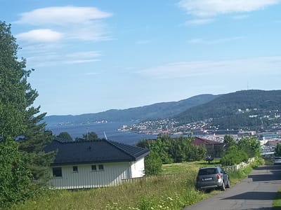 The image depicts a scenic view overlooking the city of Drammen, Norway. In the foreground, a white house with a black roof is situated on a grassy slope, with a large, leafy tree to its left. A paved road winds downhill, with a silver SUV parked on the grass beside it. In the middle ground, there are several more houses and dense green trees. The background reveals the expansive Drammen Fjord, with the city of Drammen spread out along its shores, nestled at the base of a tree-covered mountain range. The sky is a clear blue with scattered white clouds. The overall impression is one of a peaceful, sunlit day in a Norwegian coastal town.
