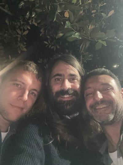 A night-time, low-angle group selfie captures three adult males positioned closely together against a dark, out-of-focus background of dense foliage. The scene is located in Pietrasanta, Italy.

From left to right:
1.  A male with short, wavy light-brown hair and minimal facial hair, wearing a dark top, faces the camera with a slight smile. The right side of his face is more illuminated than the left.
2.  A central male with long, dark-brown hair and a full dark beard and mustache, wearing a dark, ribbed knit sweater with a light-colored collar visible underneath, smiles broadly at the camera, exposing teeth. His face is brightly and uniformly lit.
3.  A male with short, dark hair and a short beard/stubble, wearing a collared shirt over a white underlayer, smiles broadly at the camera, exposing teeth. A small, dark stud is visible in his left ear. The right side of his face is more illuminated than the left.

All three individuals are looking directly towards the camera. The background consists of dark green, broad-leafed foliage, illuminated sparsely, consistent with trees or dense shrubbery in an outdoor setting under artificial lighting conditions during night hours. No other specific objects or structures are discernible in the background.