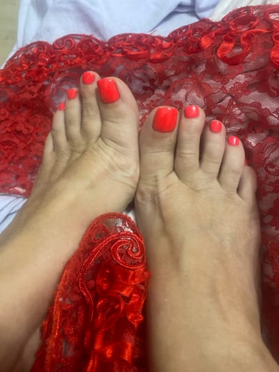 A top-down, eye-level close-up shot shows a pair of female feet adorned with vibrant orange nail polish, positioned on a white textured surface, possibly a bedsheet. The feet are positioned with the toes facing upwards and slightly spread apart, with the left foot slightly overlapping the right. A piece of red lace fabric, with intricate floral patterns and a scalloped edge, is draped over and around the feet, with a notable portion of the lace covering the arch of the left foot and extending partially under the right. The background consists of the white textured surface and more of the red lace fabric, suggesting a domestic setting. The image does not provide any information about the location in Novi Petrivtsi, Ukraine.