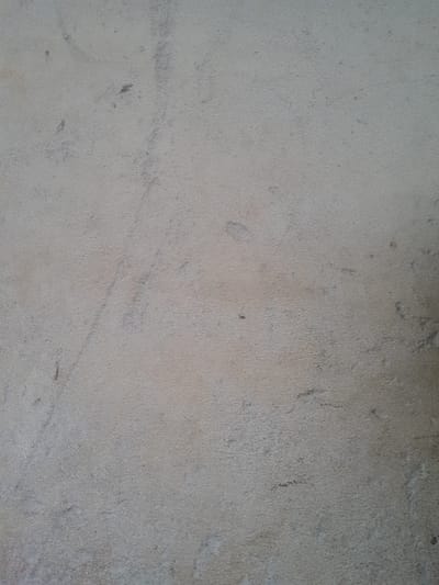 The image displays a close-up, static view of a textured surface. The surface is predominantly light grey with subtle beige and off-white undertones, exhibiting a fine-grained, irregular texture across the entire frame. Scattered across this surface are sparse, indistinct darker grey smudges, minor abrasions, and faint linear striations. There are no discernible people, specific objects, or environmental features visible that would allow for precise geographical identification within the city of Ifo, Nigeria. The scene shows no actions or interactions. The illumination appears even and diffuse.