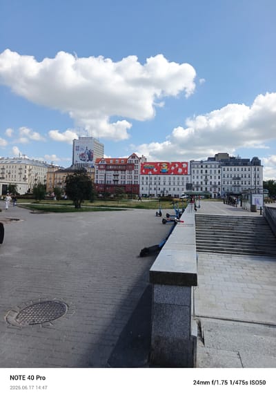 This is an outdoor photograph taken on a sunny day in Warsaw, Poland. The scene depicts a wide paved area with a prominent manhole cover in the foreground. Beyond this, a grassy area with trees separates the paved space from a row of buildings.

The buildings are a mix of architectural styles, with some featuring red-tiled roofs and ornate facades, while others are taller, modern structures. A large billboard advertising "Minionki" (Minions) with a cartoon character and multiple eyeballs is prominently displayed across the front of one of the buildings. Another billboard higher up on a taller building shows two men in suits.

Several electric scooters are parked in a line along a low stone wall. In the background, people are visible walking and interacting in the distance. To the right of the frame, a wide set of stone stairs leads up to another level of the paved area. A glass-enclosed structure, possibly a bus stop or kiosk, with the "KERASTASE" logo is also visible near the top of the stairs.

The time of day is indicated as 14:47 on June 17, 2026. The weather appears to be pleasant, with a bright blue sky dotted with fluffy white clouds. The overall atmosphere is one of a typical urban day with people going about their activities in a public space. The text visible includes "NOTE 40 Pro", "2026.06.17 14:47", "Minionki", "KERASTASE", and camera settings "24mm f/1.75 1/475s ISO50".