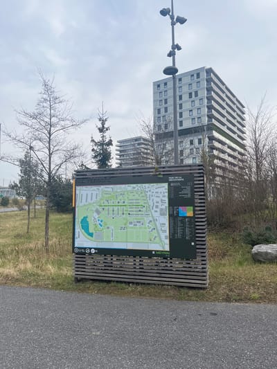 The image captures an outdoor urban or suburban scene, likely in Meyrin, Switzerland, under an overcast sky. The primary subjects are a tall, modern apartment building with numerous windows and stacked, receding balconies, and a wooden information board featuring a detailed map. Another similar building is partially visible behind the main one.

The information board stands prominently in the foreground, displaying a light green map of what appears to be a neighborhood or park, complete with roads, buildings, green areas, and a body of water. Above the map, a legend with colored squares (red, orange, green, blue) and associated text provides context. Clearly visible text on the board includes "MEYRIN" and partially visible phrases like "PARK..." and "...DÉCOUVERTE," as well as "ES VIRGIES" (likely "LES VIRGIES") along its side. Small icons and the text "R2" are also visible at the bottom of the board.

To the left of the information board, a multi-headed street light stands tall. The ground features grassy areas with patches of bare dirt, and several trees are present; some are deciduous with bare branches, suggesting late winter or early spring, while others are evergreen conifers. A large, light-colored rock is visible in the grassy background. An asphalt path or road runs along the right side of the frame. The scene is devoid of people, presenting a quiet, static view of the environment. The weather appears cloudy, as indicated by the uniformly bright, diffused sky.