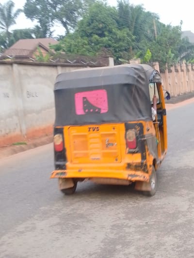 A three-wheeled yellow and black TVS auto-rickshaw, commonly known as a Keke Napep, is depicted from its rear, slightly angled right, in motion on a grey asphalt road in Alor, Nigeria. The vehicle features a black fabric soft top with a pink and black graphic sticker on the rear window. The "TVS" brand name is visible on its yellow rear panel. A person's right arm and a portion of a white shirt are visible inside the cabin on the right side. To the left, a light-colored concrete wall topped with razor wire fencing bears the graffiti "NO BILL." Behind this wall, dense green foliage and a brown-roofed building are partially visible. To the right, a brown decorative wall with pillar-like structures extends along the road, also backed by green vegetation.