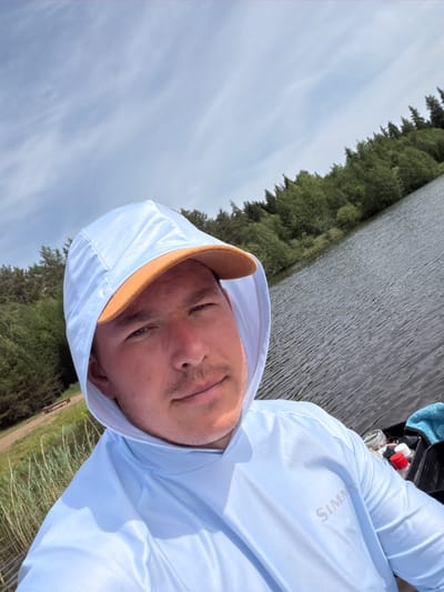 A selfie shows a person wearing a light blue hooded shirt and a baseball cap with a brown bill. The person is positioned to the right of the frame. Behind the person, a body of water extends towards the horizon on the left side of the image. Dense evergreen trees line the shore on the left side, and additional vegetation can be seen on the right side. The sky appears cloudy with patches of blue visible. Based on context, the location is likely Прикамский, Russia.