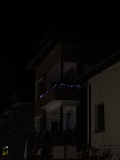The image captures a dimly lit exterior scene, likely at night, showing a portion of a multi-story building. The building has a brick facade and features a balcony adorned with colorful string lights, exhibiting blue, green, and red hues. Adjacent to this building is another structure with a lighter-colored exterior wall and a dark, narrow window. In the foreground, a metal fence and some greenery are visible, suggesting an outdoor area. The overall atmosphere is dark and slightly mysterious, with the string lights providing the only significant source of illumination. It is impossible to determine the exact location within the city of Smolyan, Bulgaria based solely on this image.
