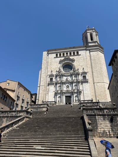 The image captures a low-angle view of a grand stone cathedral facade with a prominent rose window and intricate sculptures. A long, steep set of stone stairs leads up to the cathedral entrance, with several people ascending or standing on them. To the right of the stairs, a person holds a blue and black umbrella against a bright blue, clear sky, suggesting a sunny day. The architecture is historic and ornate, indicative of a European city. Adjacent to the cathedral and partially visible is a multi-story building with windows and balconies. The lighting indicates it is daytime, likely midday given the strong shadows. The scene appears peaceful and tourist-oriented, with people engaged in exploring the historic site. There is no visible text in the image.