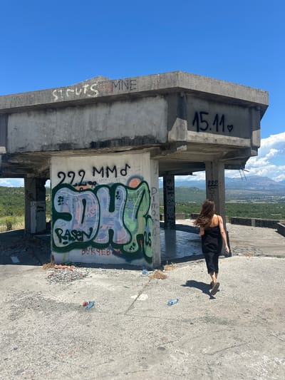 In Gornji Kokoti, Montenegro, an abandoned concrete structure, likely a former observation point or unfinished building, stands on a hill overlooking a valley. The structure is heavily graffitied. On the top horizontal concrete slab, the words "ST. CUTS MNE" are spray-painted in white, with "15.11" followed by a heart spray-painted in black on a lower section of concrete. On a vertical concrete wall, large graffiti in green and blue spray paint reads "SDH" with "RASEN" written below in smaller red letters, and "ВУКЧЕВ И" is written in red spray paint at the bottom. A woman with long brown hair, wearing a black dress and sandals, is walking away from the camera towards the right side of the frame, her shadow cast on the concrete ground. The ground is littered with debris, including broken concrete and several plastic water bottles. The background shows a lush green valley with distant mountains under a clear blue sky with scattered white clouds.