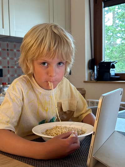 A young boy with blonde hair is sitting at a table in a home setting, in Kovačka Dolina, Montenegro. He is eating noodles from a white bowl, with a fork in his right hand. A tablet computer is positioned in front of him. The boy is wearing a yellow shirt with a white and yellow abstract pattern. In the background, there are kitchen cabinets and a window with a view of trees.