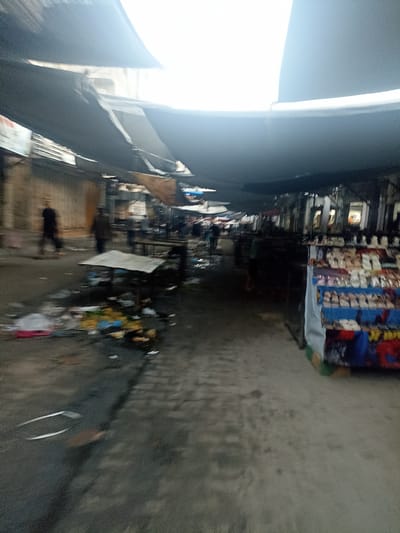 The image depicts a busy outdoor market in Gaza City, Palestinian Territories. Awnings made of dark fabric extend over the market stalls, creating a shaded walkway. The ground appears to be paved with cobblestones and is wet, with some scattered debris. Several people are visible, mostly blurred by motion, walking through the market.

On the left side of the image, a stall with a table is set up, with some items on display and more debris on the ground around it. Further down the market, there are more stalls and people.

On the right side, a stall is displaying an assortment of shoes on multiple tiers. The shoes vary in color and style. Behind this stall, other market structures and activity are visible, though less distinct. The overall impression is one of a lively, albeit somewhat disorganized, marketplace.