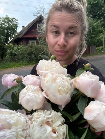 In Бронница, Russia, a young Caucasian woman with blonde hair pulled up in a bun is holding a bouquet of light pink and white peonies. She is wearing a black t-shirt and is positioned in the foreground of the image. Behind her, a weathered wooden house with a steeply pitched roof is visible, surrounded by lush green trees and bushes. The scene appears to be outdoors, on a paved surface, possibly a road or driveway, with the sky appearing overcast. Water droplets are visible on the peony petals, suggesting recent rain.