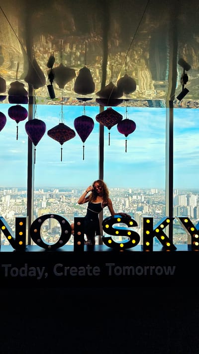A woman with curly, blonde hair and sunglasses poses for a photo in front of a panoramic city view. She is wearing a black tank top and skirt, with one hand on her hip and the other resting on a large, illuminated letter sign that spells "NOISKY". The sign is composed of individual, marquee-style letters with yellow lights. Above the sign, several ornate, Asian-style lanterns hang from the ceiling, which reflects the city lights and the lanterns themselves. The woman appears to be cheerful and relaxed.
The backdrop is a cityscape with numerous buildings stretching to the horizon, suggesting a large, urban environment. The sky is a clear blue with scattered clouds, indicating daytime. The overall scene suggests the photo was taken at a high vantage point, likely a skyscraper observation deck or a rooftop bar.
Below the "NOISKY" sign, a second, darker sign reads "Today, Create Tomorrow" in grey, block lettering. The presence of the "NOISKY" sign and the cityscape of Hanoi strongly suggests the location is in Hanoi, Vietnam. The time of day appears to be late afternoon or early evening, as the sky is bright but the city lights are beginning to twinkle.