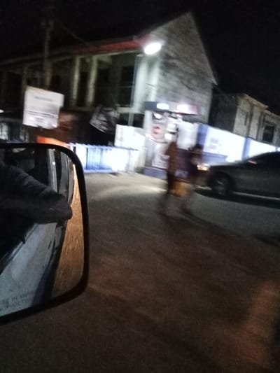A blurry, low-light photograph captures a nighttime street scene in Orie Uratta, Nigeria. The image is taken from a moving vehicle, with the driver's side mirror occupying the lower-left foreground, reflecting a partially visible arm extending from the vehicle's interior. In the background, multiple multi-story buildings with dark windows line the street. One building is partially illuminated by a bright, circular overhead light fixture. Several white, rectangular signs or posters, indistinct due to blur, are affixed to poles or building facades. Two highly blurred human figures appear to be walking on the right side of the road or sidewalk in the middle ground, their features indistinguishable. A dark-colored passenger vehicle is partially visible on the far right, also blurred by motion. A line of light-colored barriers or fences is present along the roadside in front of the buildings. The asphalt road surface displays reflections from ambient lighting.