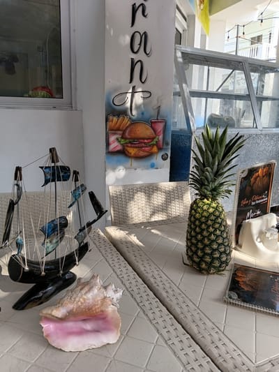 The image captures an outdoor scene, likely a cafe or restaurant patio, given the presence of white wicker furniture and a tiled tabletop. Sunlight suggests it's daytime. A pineapple rests on the table, positioned near a menu with "Rincón Bar Restaurant" visible. Next to the pineapple are several decorative items: a large conch shell with a pink interior, a dark, horn-shaped object, and a mobile featuring small, colorful fish suspended from strings attached to wooden dowels.
In the background, a glass-paneled structure, possibly a walkway or part of a building, allows light to filter through, illuminating the scene. A mural or graffiti is visible on a white wall, depicting a hamburger and fries, and stylized lettering that appears to be "La..."
The overall impression is a relaxed, casual setting, possibly in a tropical or coastal environment, indicated by the pineapple, conch shell, and fish mobile. There are no people visible in the image.
