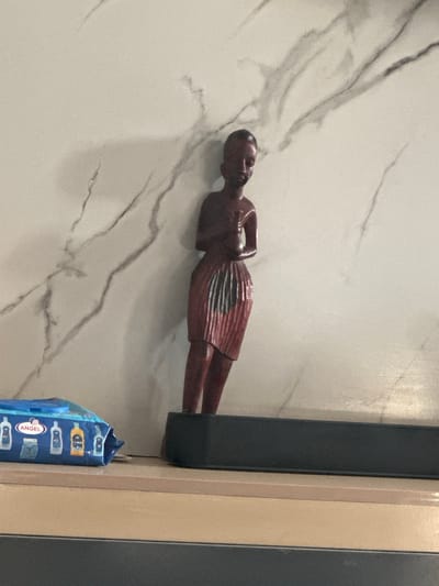 A wooden figurine of a person is displayed horizontally against a white, marbled surface. The figurine is dark brown and appears to be wearing a patterned skirt. To the right of the figurine, a black object, possibly a speaker or soundbar, stands vertically. Next to that, a beige vertical beam is visible. At the bottom right of the image, a blue package with various colorful icons and the word "ANGEL" in a pink circle is partially visible. The scene is indoors, with artificial lighting. There are no indications of time of day or weather conditions. The setting appears to be a home or a display area. The figurine itself is the main subject, with the surrounding objects serving as context.