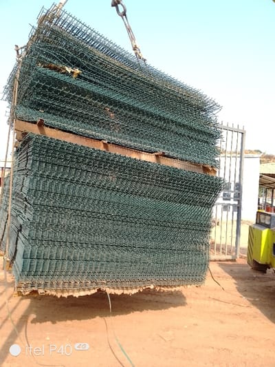 This image, captured in the city of Hoche, Nigeria, presents a large, vertically stacked bundle of approximately 25-30 layers of green-coated welded wire mesh panels in an outdoor setting. The rectangular mesh panels are stacked on what appear to be wooden pallets or support beams. A heavy-duty metal hook, attached to chains or cables, is visible at the top center, engaged with the uppermost section of the stack, indicating the bundle is either being lifted or lowered by an unseen crane. The ground beneath is reddish-brown dirt. In the bottom foreground, two thin, dark cables or hoses lie on the ground. To the right, a yellow and black forklift is partially visible. Behind the forklift, a grey metal gate is present, and further in the background, a white building with a brown roof structure can be seen. The sky above is clear and light blue. No individuals are visible in the scene.