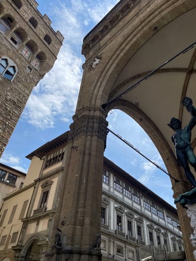 The image is a low-angle shot in Florence, Italy, showcasing a view upwards through the archway of the Loggia dei Lanzi, with the bronze statue of Perseus with the Head of Medusa visible. The archway is constructed of weathered stone, with visible detailing and supports. A building with a light-colored facade, many windows, and elaborate trim is situated to the right of the arch. Another stone building is to the lower left, exhibiting arched windows and decorative shield insignias. The sky is visible through the space framed by the archway, displaying light blue sky and some scattered white clouds.