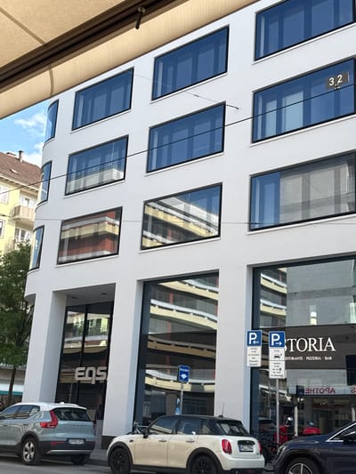 A contemporary white building with large, dark-framed windows is the primary subject. The windows reflect the surrounding environment, including other buildings and the sky. On one of the upper windows, the numbers "3.2" are visible. Below, at street level, the building features large glass storefronts. One storefront has the white logo "eqs" displayed prominently. Next to this, another storefront displays the white text "TORIA" with smaller text below that reads "RISTORANTE PIZZERIA BAR".
In front of the building, two cars are parked. The car on the left is a light blue Volvo SUV with license plate "L R KS 1933". The car on the right is a white Mini Cooper with license plate "M LL 7994". Two blue parking signs are visible on poles between the cars and the building.
The scene appears to be in a city, likely a commercial area given the storefronts and parking signs. The presence of the German license plate and the text on the signs indicate the location is Germany. The architectural style of the building suggests a modern urban environment.