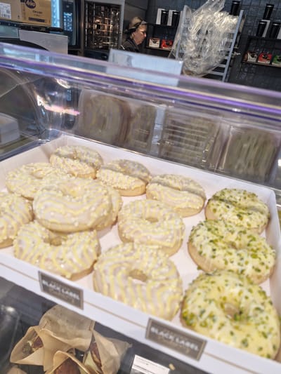 The image shows a close-up view of donuts displayed in a bakery case. The donuts have a light-colored glaze and are decorated with yellow icing drizzles and some with green sprinkles, possibly pistachios. There are two distinct types of donuts visible, one with yellow drizzle and another with green sprinkles. A label that reads "BLACK LABEL" is visible at the bottom of the display case. In the background, behind the display case, a person wearing glasses and a dark headscarf is partially visible, looking towards the left. Behind the person, there are shelves with packaged items and what appears to be kitchen equipment, including a large oven. The overall scene suggests a retail environment within a food establishment. No specific location in Bremen, Germany, can be determined from the image.