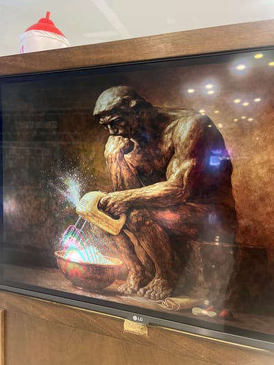 The image shows a digital display, likely a television, with a wooden frame. The screen displays an image of a human-like form huddled on the ground in a fetal position. The figure has a skull-like head. A kitchen hand mixer is positioned near the figure, appearing to be mixing something into a bowl. The artwork has brown tones throughout. The location is not apparent from the image.