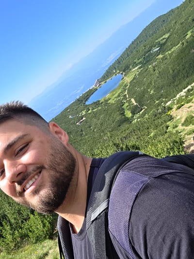 A young man with short dark hair and a beard smiles at the camera in a selfie taken outdoors. He is wearing a dark blue t-shirt and a dark backpack with grey straps. The background shows a steep, tree-covered mountainside and two lakes. The sun is shining, and the sky is clear blue. The location is Rila National Park in Bulgaria, which is near the city of Bansko. The lakes visible are the Beli Iskŭr Lakes, also known as the Seven Rila Lakes.