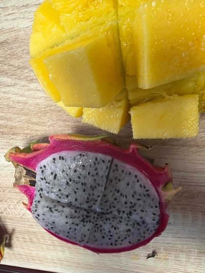 The image shows a close-up, overhead shot of chopped mango and a halved dragon fruit resting on a light-colored wooden surface. The mango pieces are a bright yellow and appear juicy, with some slight glistening. The dragon fruit is cut lengthwise, revealing its white flesh speckled with small black seeds, enclosed by a vibrant pink skin with green tips. The wooden surface has a visible grain pattern. No people or actions are depicted, and the setting is not identifiable as being in Męcina, Poland.