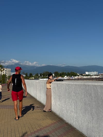 A man and a woman are captured in a vertical outdoor shot on a sunny day. The man, wearing a black Reebok t-shirt and red shorts, stands on a brick pathway, looking towards the left. He has a red cap on and tattoos on his arms. The woman, in a light-colored top and beige pants, leans against a textured concrete wall, holding a black bag. She is looking upwards.
In the background, a cityscape with white buildings is nestled against a backdrop of lush green trees and a majestic, blue mountain range under a clear, bright sky. The sun casts distinct shadows on the pathway, suggesting it is either morning or late afternoon. The overall atmosphere appears relaxed and casual, perhaps during a sightseeing excursion. The word "Reebok" is visible on the man's t-shirt. The scene is set in Sirius, Russia.