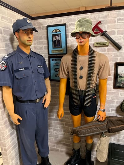 Two mannequins stand against a brick wall. One wears a khaki t-shirt, black shorts, ammo belts, glasses, and a hat, with black boots completing the outfit. The other mannequin is dressed in an air force uniform with a cap and black boots. The wall is adorned with framed pictures and tools, adding to the display.