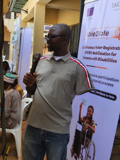 The image depicts an outdoor event likely taking place in Maiburiji, Nigeria. A man wearing glasses and a grey polo shirt with red, white, and blue stripes on the sleeves is speaking into a microphone. He is wearing blue jeans. In the background, a large banner is visible with text that includes "Able2Vote," "Continuous Voter Registration (CVR) Mobilization for Persons with Disabilities," and "Inclusive participation in the electoral process." The banner also features an image of a smiling woman in a wheelchair holding a voter's card, and the European Union logo.
Several chairs are arranged in front of the speaker, with some people seated on them, also appearing to be listening to the speaker. The overall setting appears to be a public gathering, possibly a community meeting or rally, focused on voter registration for people with disabilities. The lighting suggests it is daytime. Other visible text on a smaller banner in the background says "NO E" and "RE." The atmosphere appears to be engaged and informative, given the subject matter and the speaker's interaction with the microphone.