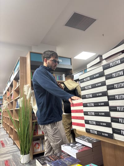 The image depicts the interior of a bookstore in Prague, Czechia. A male with dark hair and a beard, wearing sunglasses, a dark blue quarter-zip fleece, and light tan cargo pants, stands in the foreground, facing right. His right hand rests on a stack of books with red and white striped spines, titled "CAUGHT UP BY NAVESSA ALLA." Behind him and slightly to the right, a second male is partially visible from the back, wearing a beige hooded sweatshirt and a black backpack. To the right of the foreground subject, a large display table holds multiple stacks of books, including numerous copies of "PHANTOM" by R.R. CARLTON with black covers, and other books like "SWEET RUIN" and "RUTHLESS CREATURES." In the midground, wooden bookshelves line the walls, filled with various titles, some sections labeled "NON FICTION" in teal. A tall, potted artificial plant with green blades and white feathery plumes is situated on the patterned carpeted floor to the left. The ceiling is white with visible square ventilation grilles and recessed lighting fixtures, providing ample illumination to the scene.