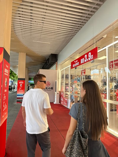 In the Yuexiu District of China, a man wearing a white t-shirt and sunglasses is walking on a red carpet next to a store with a glass facade. He is facing away from the camera, looking towards the store. A woman with long brown hair, carrying a leopard-print bag, is walking ahead of him on the same red carpet, also facing the store. The store's interior is visible through the glass, displaying clothing and bags on shelves. Red banners with Chinese text and numbers are displayed on the store's windows, indicating a sale. Other people and buildings are visible in the background, suggesting a bustling urban environment. The overhead structure is made of light-colored wooden slats.