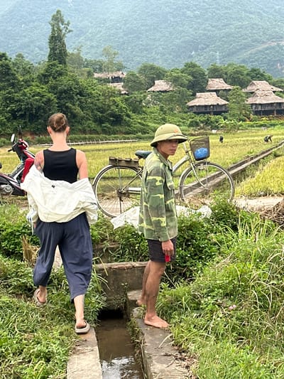 In Mai Chau, Vietnam, a man wearing a camouflage shirt and hat stands near a rice paddy. He is barefoot and holding a red object. Next to him, a woman with tattoos on her arm walks away from the camera, carrying a white jacket. A bicycle is parked behind them, with a blue basket on the handlebars. In the background, traditional stilt houses with thatched roofs are nestled among lush green trees, with rolling hills in the distance. A motorcycle is parked to the left of the woman.