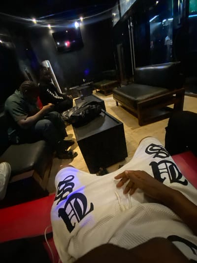 The image depicts an indoor setting in Iranla, Nigeria, possibly a lounge or entertainment venue. Two people are visible, one wearing a green shirt and another wearing a black shirt with white letters, both seated in upholstered chairs. A person wearing white shorts adorned with large black lettering sits on a red cushioned bench. In the background, there is a wall-mounted television and ambient lighting. Additional furniture includes wooden chairs with dark upholstery.