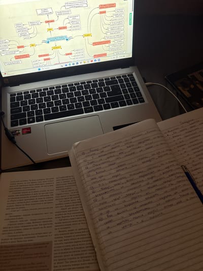 An indoor scene depicts a study or work setup on a dark, textured surface, possibly a bed or fabric cover, under ambient dim lighting. A silver Acer laptop is open on the left, displaying a complex, multi-colored mind map on its screen. The mind map, written in Bulgarian, centers on "Героите в повестта „Крадецът на праскови“, Емилиян Станев" (The Heroes in the novella "The Peach Thief", Emilian Stanev), with nodes detailing characters like "Елисавета" (Elisaveta), "Иво" (Ivo), and "полковникът" (the colonel), and attributes such as "описание" (description) and "характер" (character). The laptop screen also shows a weather widget indicating "21°C Mostly cloudy" and a system clock displaying "23:28" and a date of "7.4.20XX". The laptop's keyboard features a QWERTY layout with Cyrillic sub-labels, and stickers for AMD Ryzen and AMD Radeon Graphics are visible on the left palm rest. A black charger cable is plugged into the left side of the laptop. To the right of the laptop, an open, lined spiral notebook lies flat, filled with handwritten notes in blue ink, also in Bulgarian, appearing to be analytical summaries. A blue ballpoint pen rests diagonally across the notebook. An open physical book, also with Bulgarian text, is partially visible beneath the notebook and to the left. A dark, patterned object is partially visible on the far right. The image contains no visual information to confirm a location in the city of Lom, Bulgaria.