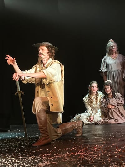 The scene depicts a theatrical performance taking place on a stage with a stark black backdrop in Novoshakhtinsk, Russia. In the foreground, a male actor, characterized by a long mustache and goatee, a wide-brimmed hat, light-colored shirt, tan tunic, and dark knee-high boots, kneels on his right knee. His left hand rests on the pommel of an upright sword, its tip on the confetti-strewn dark stage floor, while his right arm is raised and extended in a dramatic gesture. His gaze is directed off-frame to the left. To his right, two young females, dressed in period-style long dresses (one off-white, one light brown), sit on the stage, observing the male figure with serious expressions. Behind them, an elderly woman with light grey hair, wearing a loose grey dress and a necklace, stands partially visible, also looking towards the central male figure. The stage floor is dark, scattered with numerous small, light-colored paper fragments or confetti, illuminated by focused stage lighting.